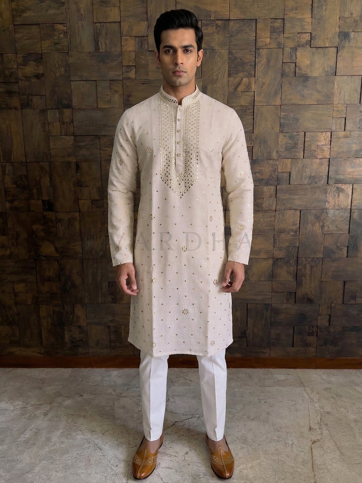 Man wearing a cream kurta with white pants against a wooden paneled wall. Cream khadi cotton kurta with gold floral mukaish work for men paired with white churidar and brown mojari