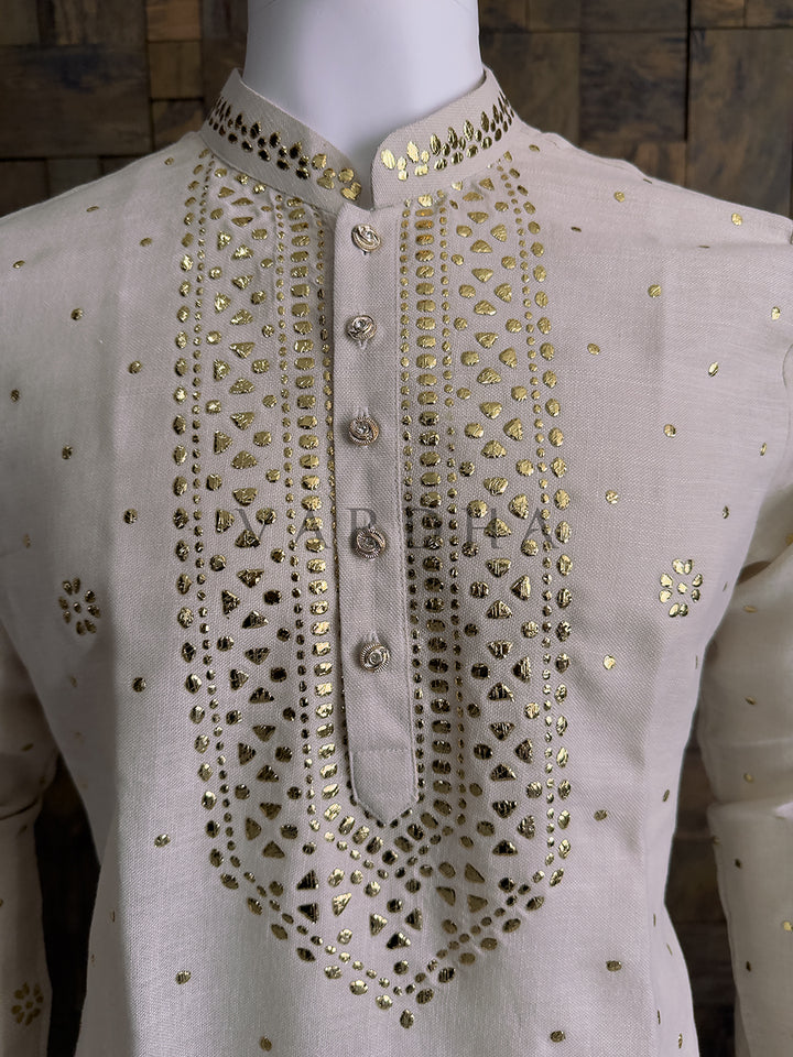 Mannequin wearing a light-colored shirt with gold embellishments on a wooden background. Classic cream ethnic kurta in khadi cotton with gold mukaish embellishments and straight fit silhouette