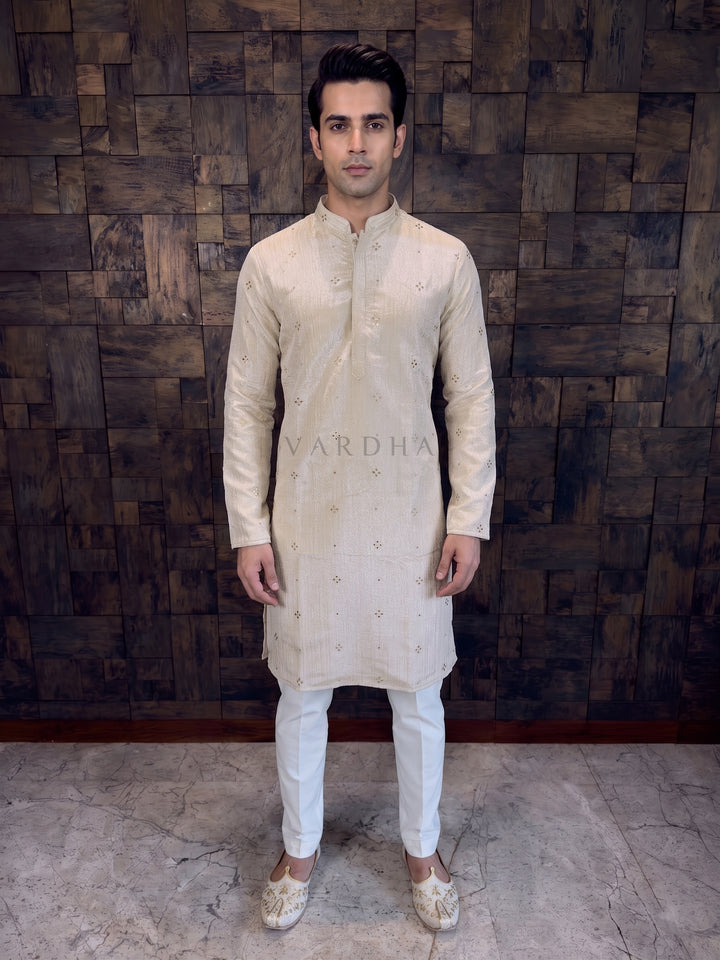 Man wearing a cream kurta with white pants against a wooden paneled wall. Viscose Tissue Crush fabric, the kurta features a permanent crinkled finish that adds depth and an excellent drape. The rich cream base is delicately embellished with scattered sequin&amp;nbsp;mukaish tiny