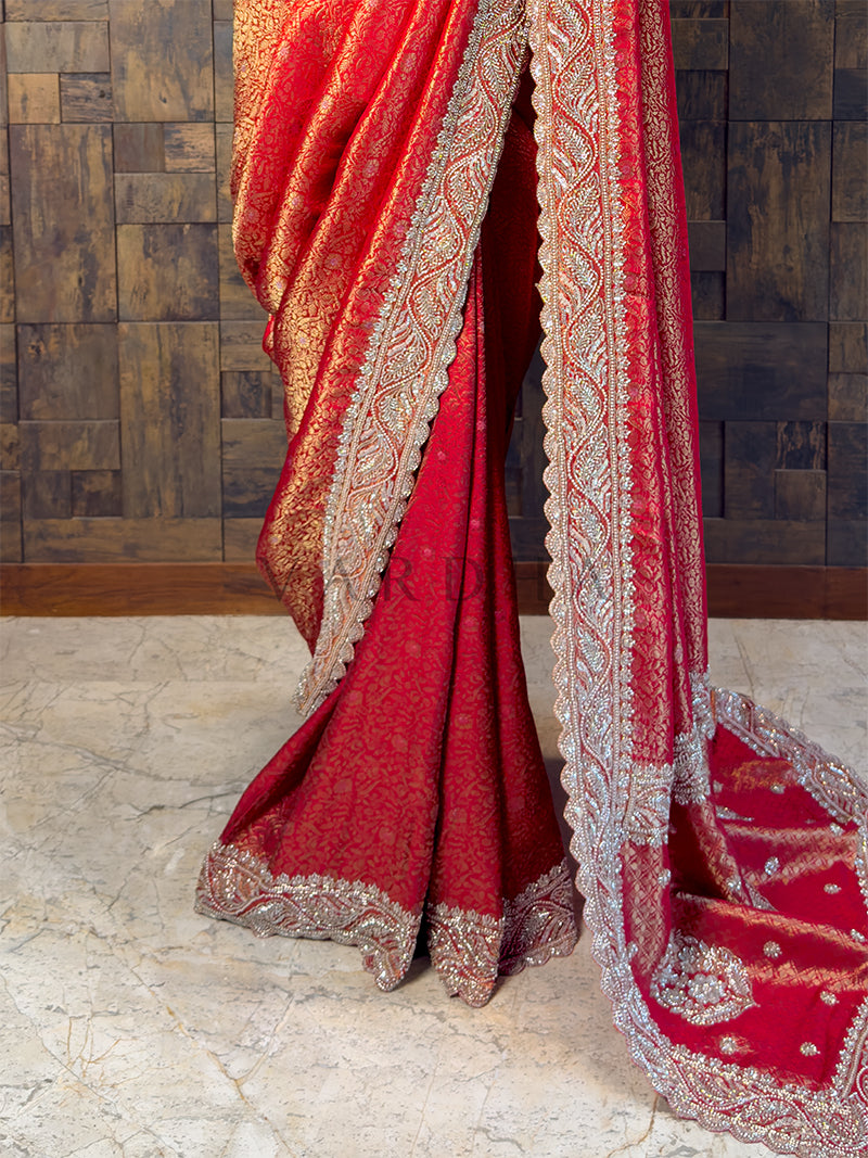 Red saree with intricate gold patterns on a stone floor and wooden wall background. Heavy Gold Zari Jaal and Stone Embellished Pallu on Red Wedding Saree