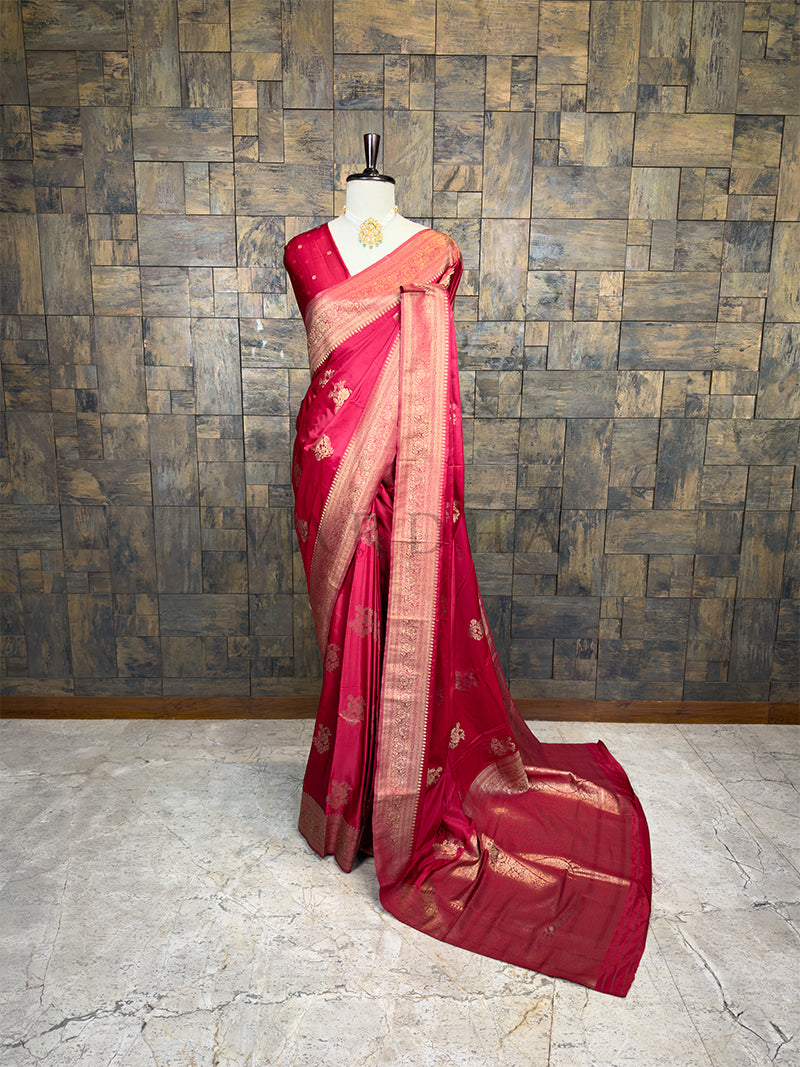 Red saree with gold patterns on a mannequin against a textured wall. House of Vardha Crimson Red Satin Saree featuring Light Gold Zari Floral Motifs and a matching border, elegantly draped on a mannequin. We offer Ready to wear, Pre drape service, Saree Add on, Custom Stitched Blouse, Fall and Edging, Gift Packaging at Complimentary Price!