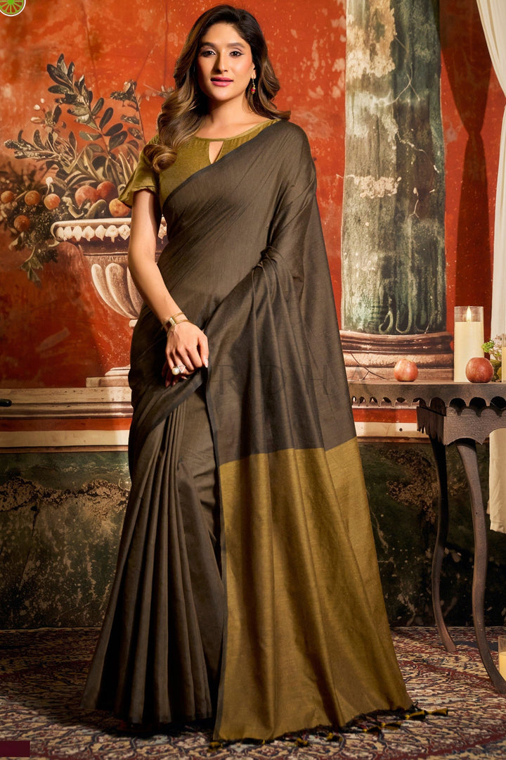 Woman wearing a gray and green saree in an indoor setting with decorative elements. Full front view of a woman wearing an elegant dark brown mal cotton saree featuring a khaki mustard contrast pallu and fancy tassels.