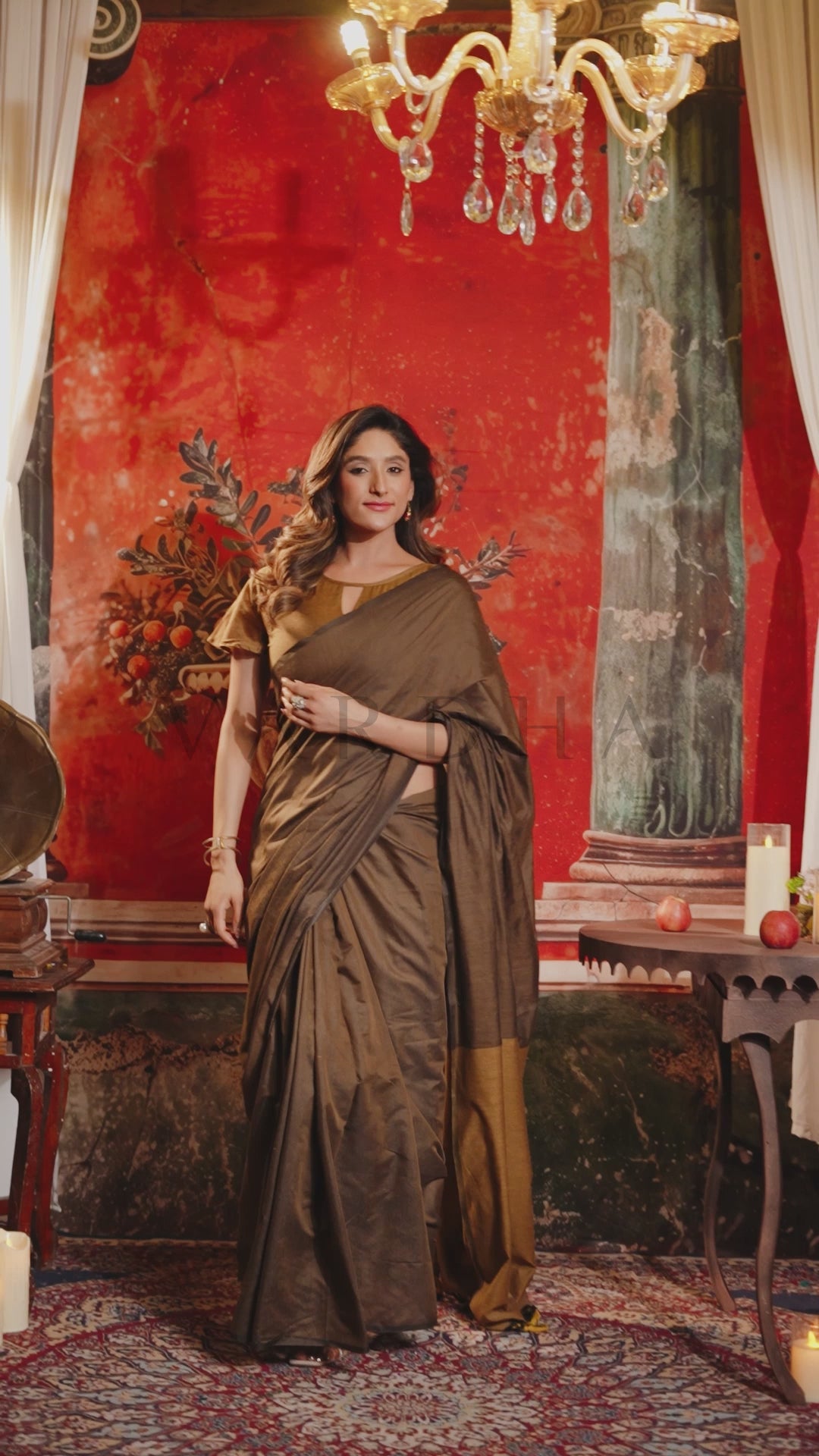 A woman wearing a brown silk saree with a gold embroidered yoke stands in an ornate room with a red wall, chandelier, and patterned rug. Stylish front drape of the lightweight dark brown mal cotton saree, showcasing the earthy dual-tone look for casual cultural outings.