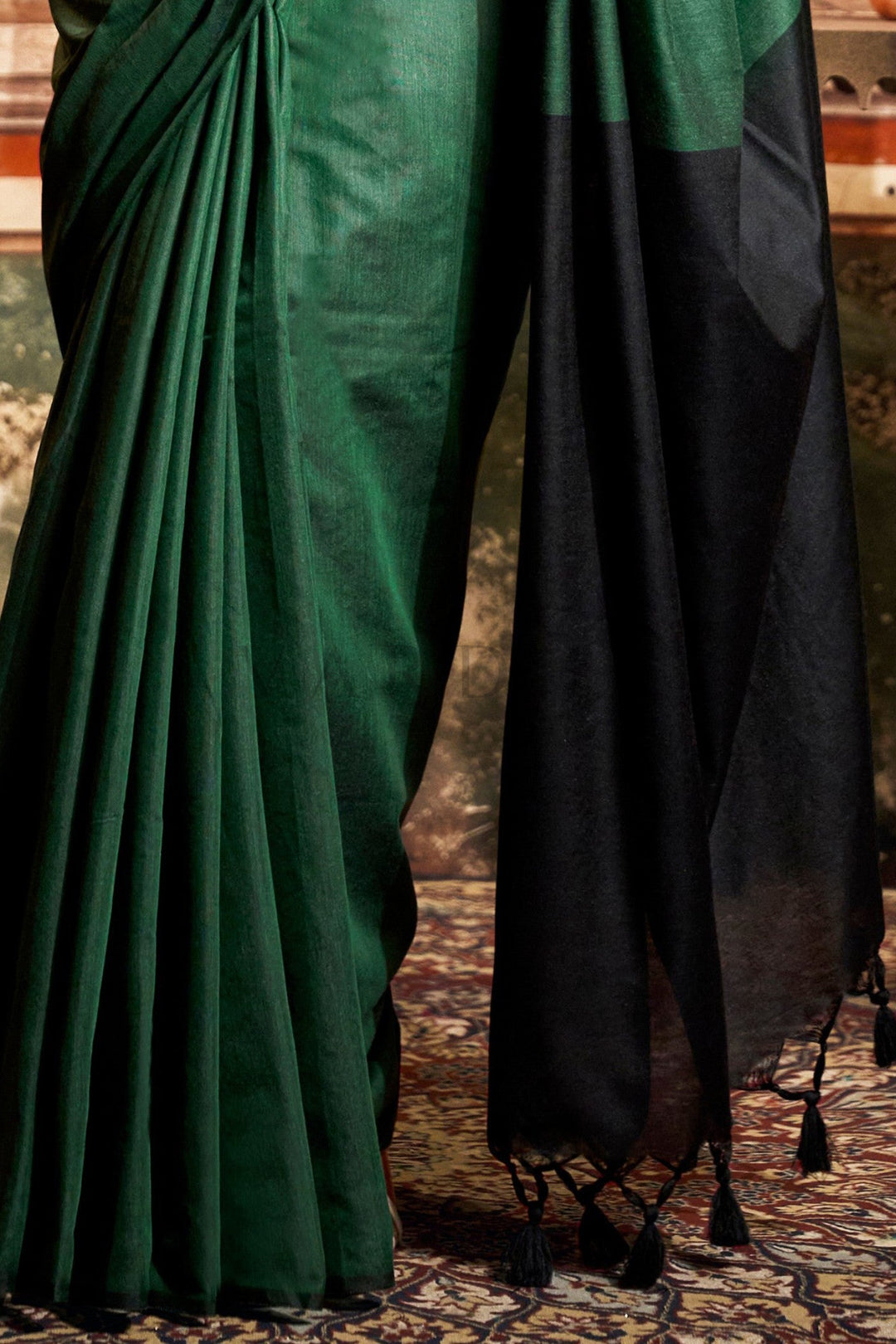 A close-up of a dark green and black saree, showing the pleated green body, the black pallu with tassel ends, and the draped fabric. Close-up showcasing the striking solid black contrast pallu and beautiful fancy black tassels on the hem of the green mal cotton saree.