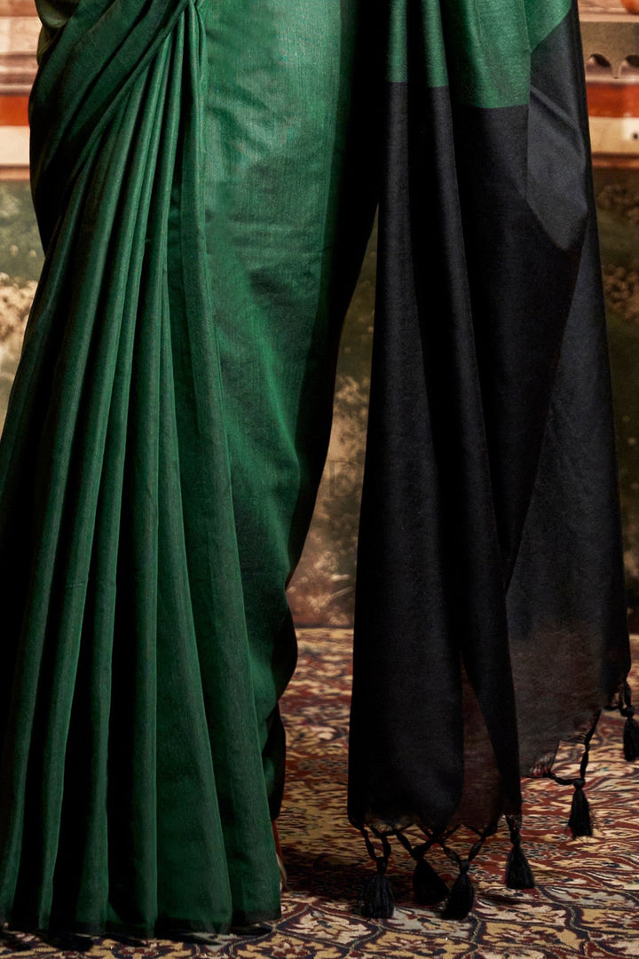 A close-up of a dark green and black saree, showing the pleated green body, the black pallu with tassel ends, and the draped fabric. Close-up showcasing the striking solid black contrast pallu and beautiful fancy black tassels on the hem of the green mal cotton saree.