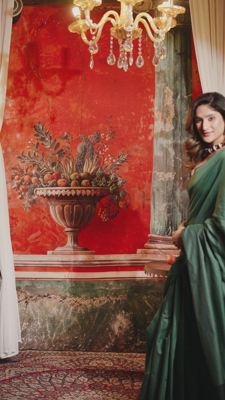 A woman in a flowing dark green gown stands beside a large red wall mural featuring a classical urn with fruit and flowers, with a chandelier hanging above Beautiful woman modeling a lightweight dark green plain mal cotton saree in a warm, festive vintage setting.