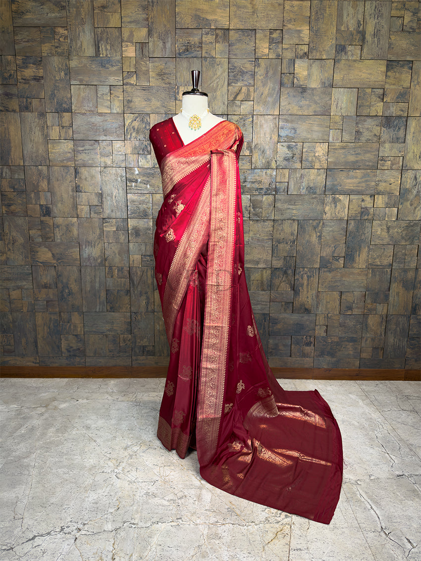 Red saree with gold patterns on a mannequin against a textured wall. House of Vardha Deep Maroon Satin Saree featuring Gold Zari Floral Motifs and a matching border, elegantly draped on a mannequin. We offer Ready to wear, Pre drape service, Saree Add on, Custom Stitched Blouse, Fall and Edging, Gift Packaging at Complimentary Price!