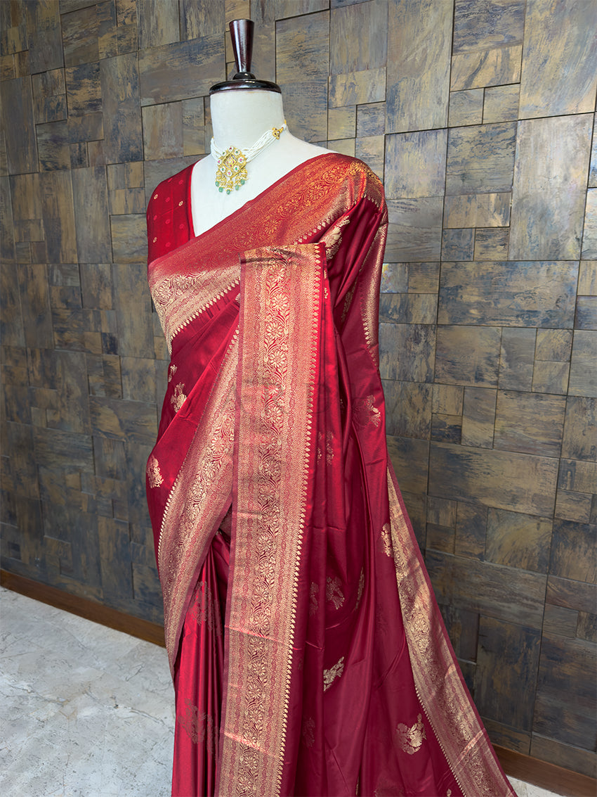 Red saree with gold patterns on a mannequin against a textured wall. Close-up view of the House of Vardha Deep Maroon Satin Saree highlighting the detailed Gold Zari floral motifs and the heavy woven border. We offer Ready to wear, Pre drape service, Saree Add on, Custom Stitched Blouse, Fall and Edging, Gift Packaging at Complimentary Price!