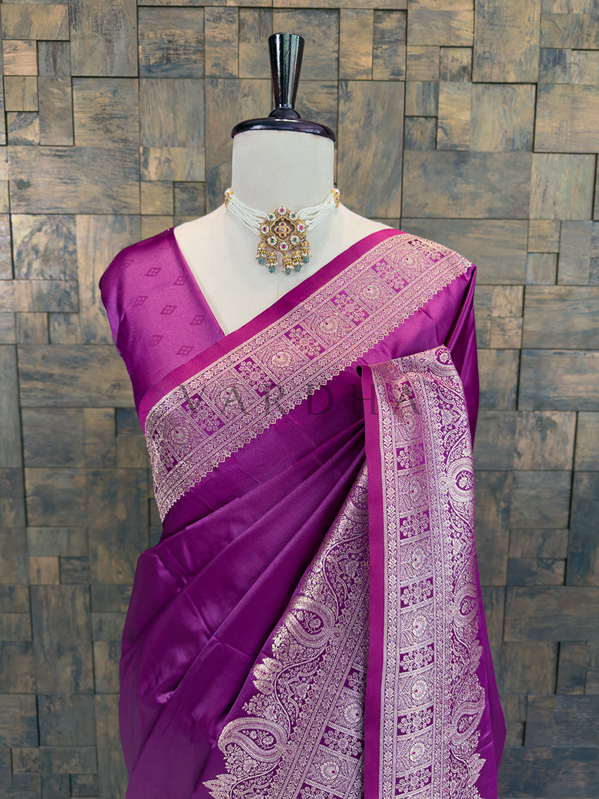 Purple saree with white patterns on a mannequin against a stone wall background Close-up of a deep purple woven satin ethnic saree styled on a mannequin display, paired with a traditional pearl choker necklace.