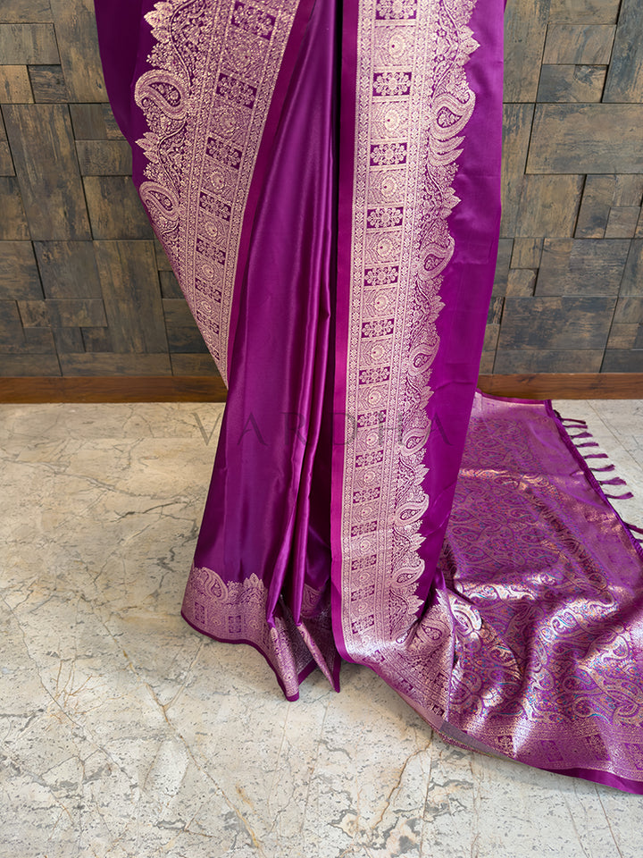 Purple saree with white intricate border on a stone floor and wooden wall  background Lower pleats and fluid drape of a glossy violet Banarasi-style satin silk saree showcasing the broad paisley border. Detailed floor view of the rich, densely woven golden zari pallu with matching tassels on the luxurious purple satin saree.