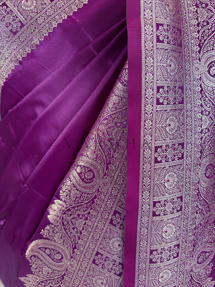 Purple saree with intricate white patterns Close-up texture shot highlighting the solid glossy purple fabric contrasting beautifully with the intricate floral zari border. Elegant party wear deep purple satin silk saree highlighting its smooth drape, lustrous shine, and exceptional zari craftsmanship.