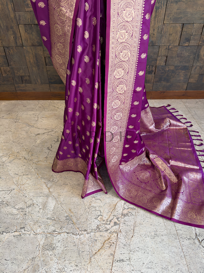 Purple saree with gold patterns on a stone floor Lower pleats and fluid drape of a glossy plum Banarasi-style satin silk saree showcasing the broad circular floral border.