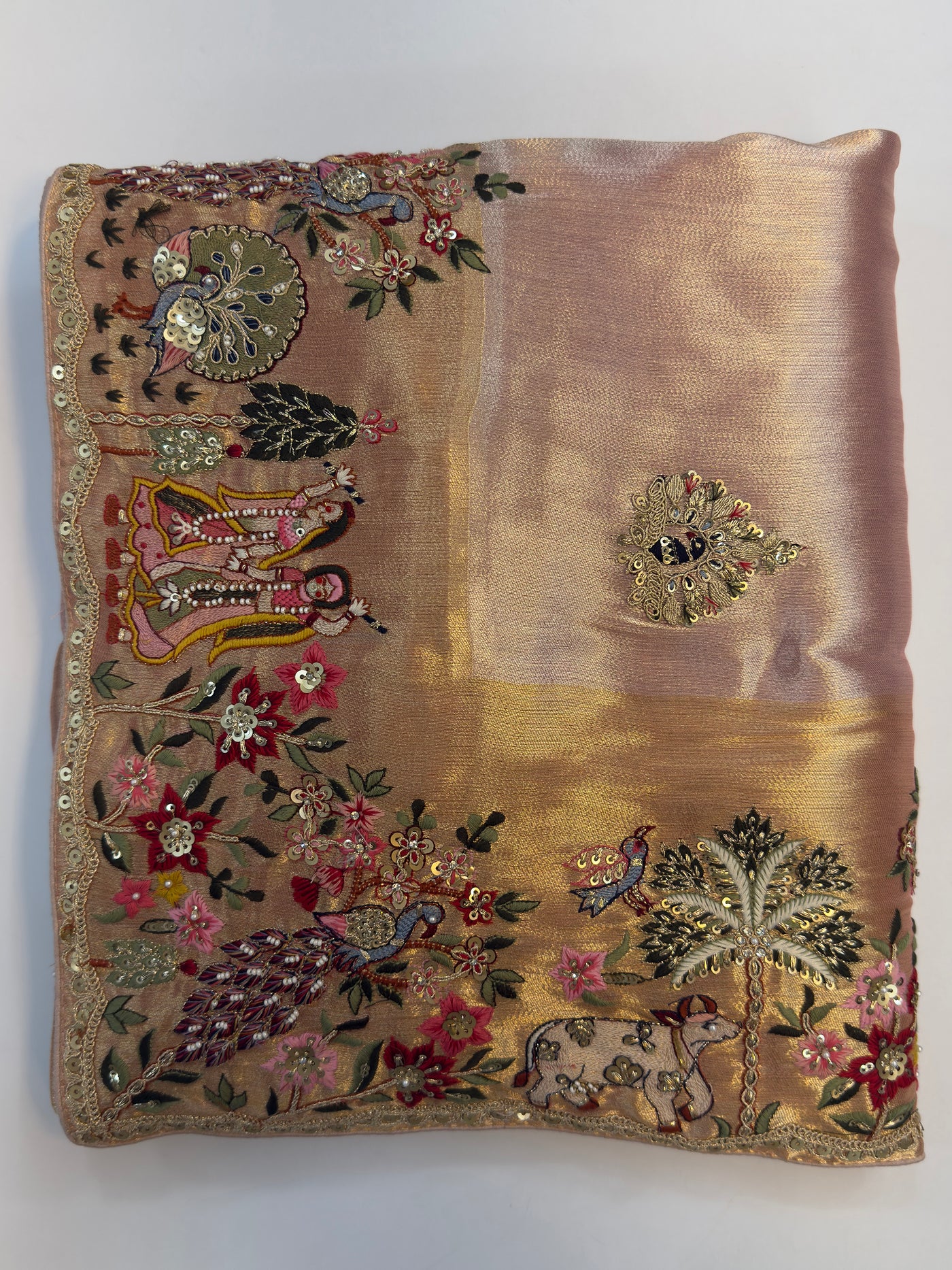Hand-embroidered pink–lilac tissue silk saree with Radha Krishna and floral motifs captured in folded view showcasing luminous fabric glow.

