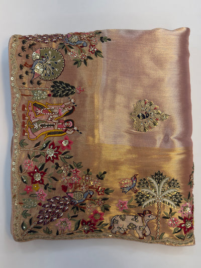 Hand-embroidered pink–lilac tissue silk saree with Radha Krishna and floral motifs captured in folded view showcasing luminous fabric glow.

