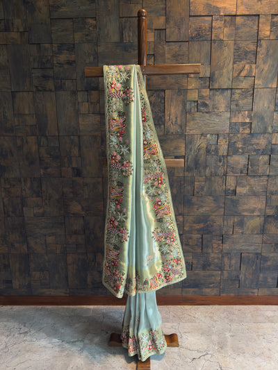 Pista green tissue art silk saree with gold undertone, hand embroidery, mirror work and zari, featuring Radha Krishna, peacocks, cows and floral trees on an ornate story border.
