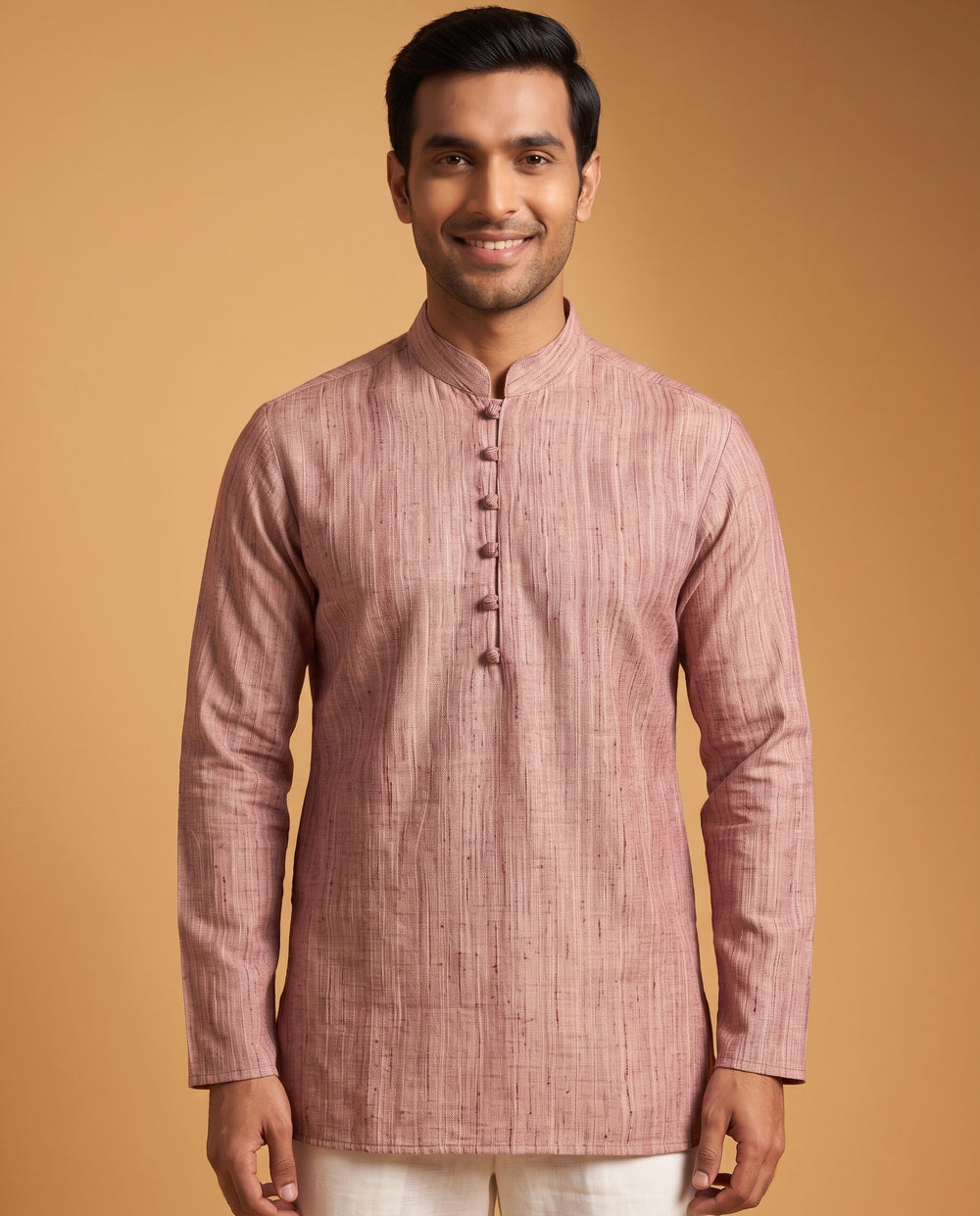 A man wearing a dusty rose pink textured kurta with a mandarin collar and buttoned front, paired with off-white trousers, set against a solid warm tan background. Close-up shot of a dusty cedar textured cotton short kurta featuring a mandarin collar and traditional button detail.