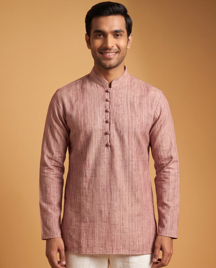 A man wearing a dusty rose pink textured kurta with a mandarin collar and buttoned front, paired with off-white trousers, set against a solid warm tan background. Close-up shot of a dusty cedar textured cotton short kurta featuring a mandarin collar and traditional button detail.