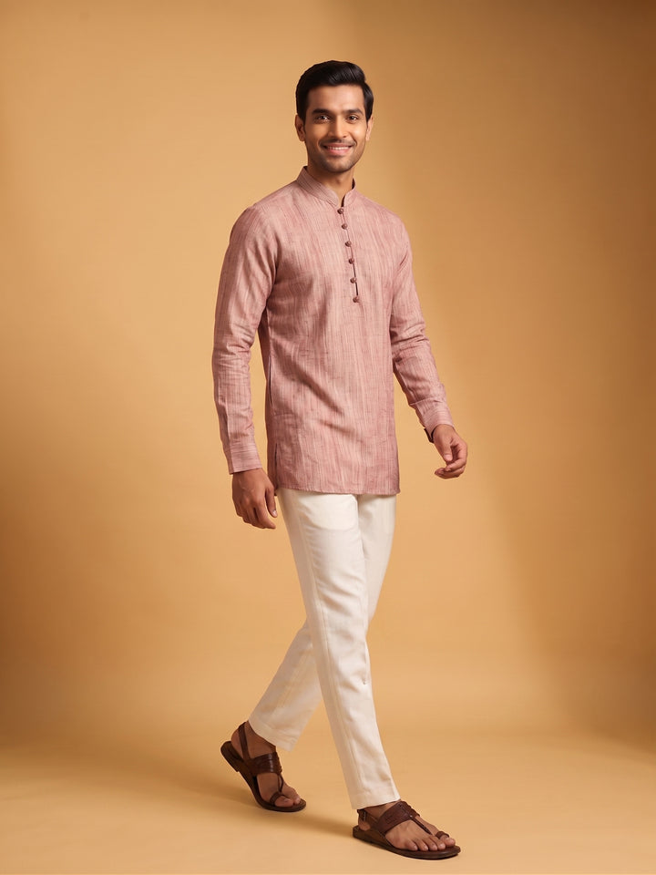 A man modeling a light pink long-sleeve kurta with white trousers and brown sandals, standing against a solid warm beige studio background A man walking in a dusty cedar textured cotton short kurta and white trousers, side-angle view showcasing fit.