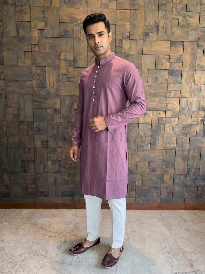 Man wearing a mauve kurta with white pants against a wooden paneled wall. Men’s Dusty Mauve Pink cotton kurta featuring a woven vertical micro-stripe dobby texture that adds visual depth and elongates the frame.