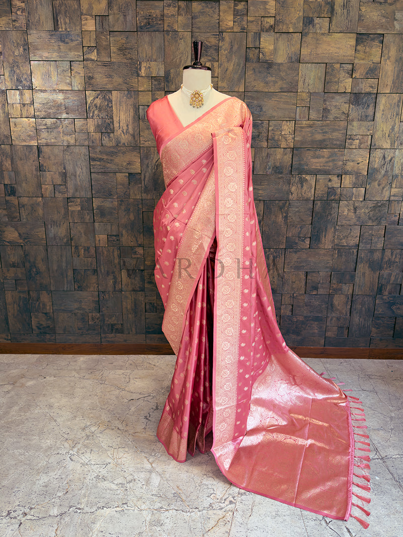 Pink saree with a mannequin against a textured wall. Full front display of a dusty rose pink satin silk saree featuring golden zari paisley bootis draped gracefully on a mannequin. Elegant party wear soft pink satin silk saree highlighting its smooth drape, lustrous shine, and exceptional zari craftsmanship.