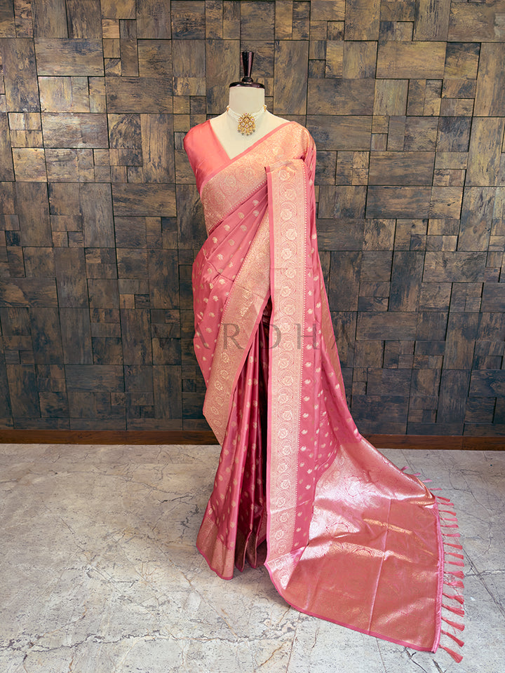 Pink saree with a mannequin against a textured wall. Full front display of a dusty rose pink satin silk saree featuring golden zari paisley bootis draped gracefully on a mannequin. Elegant party wear soft pink satin silk saree highlighting its smooth drape, lustrous shine, and exceptional zari craftsmanship.