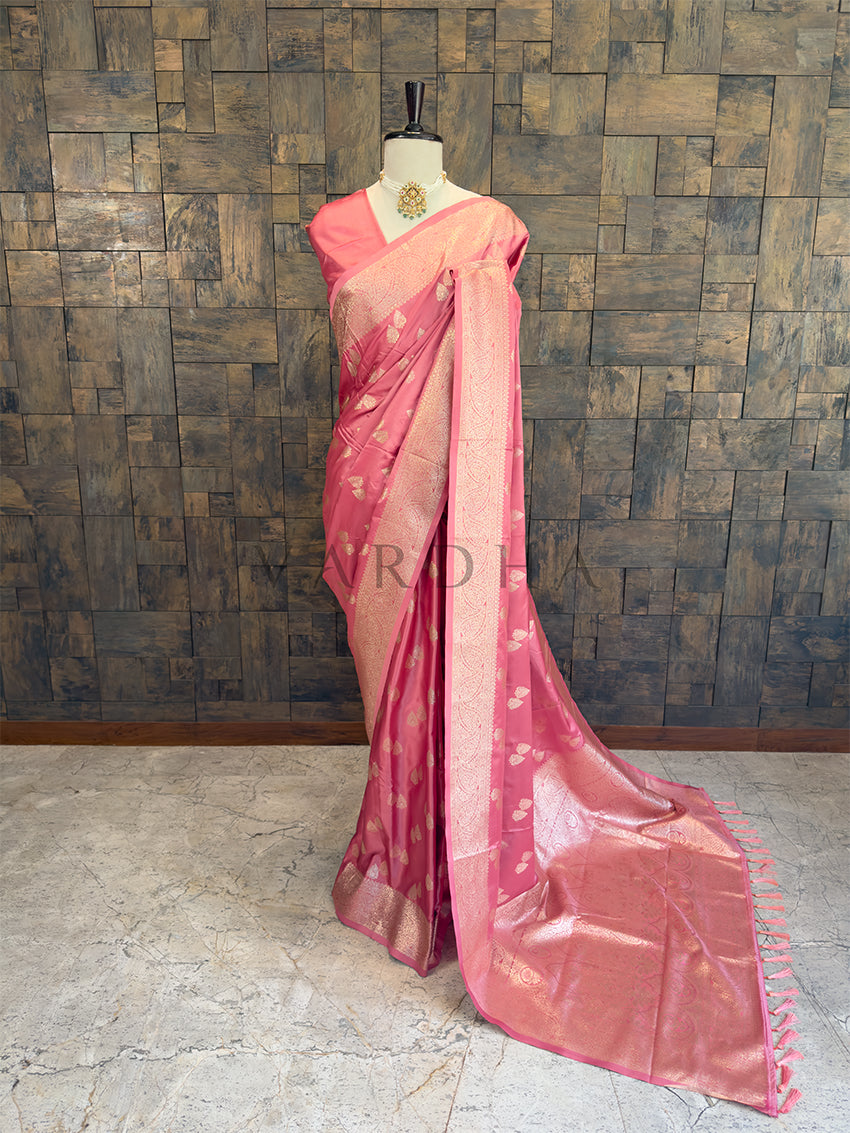 Pink saree with a subtle pattern on a mannequin against a textured wall. Dusty Rose Satin Silk Banarasi Saree with Zari Woven Booti & Heavy Pallu Full front display of a dusty rose pink satin silk saree featuring golden zari bootis draped gracefully on a mannequin.