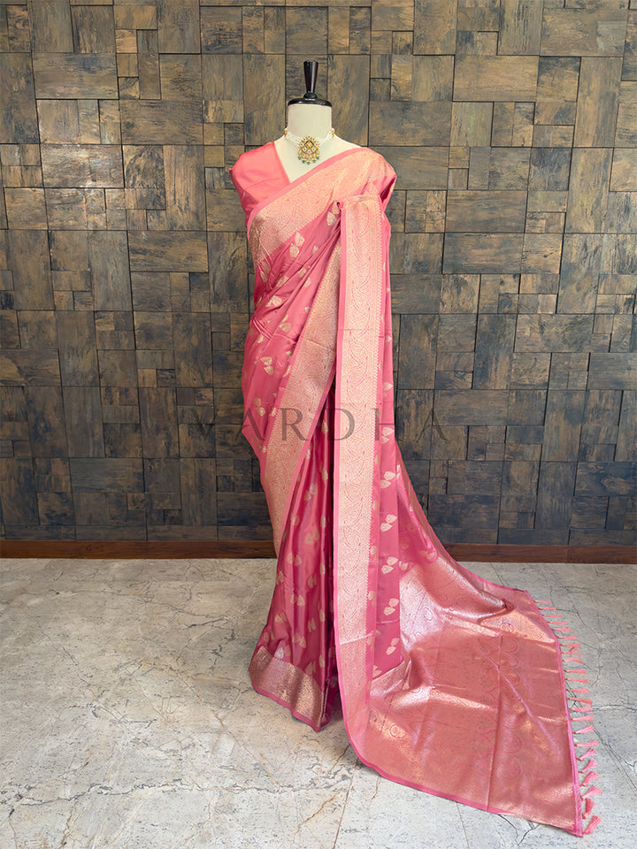 Pink saree with a subtle pattern on a mannequin against a textured wall. Dusty Rose Satin Silk Banarasi Saree with Zari Woven Booti &amp; Heavy Pallu Full front display of a dusty rose pink satin silk saree featuring golden zari bootis draped gracefully on a mannequin.