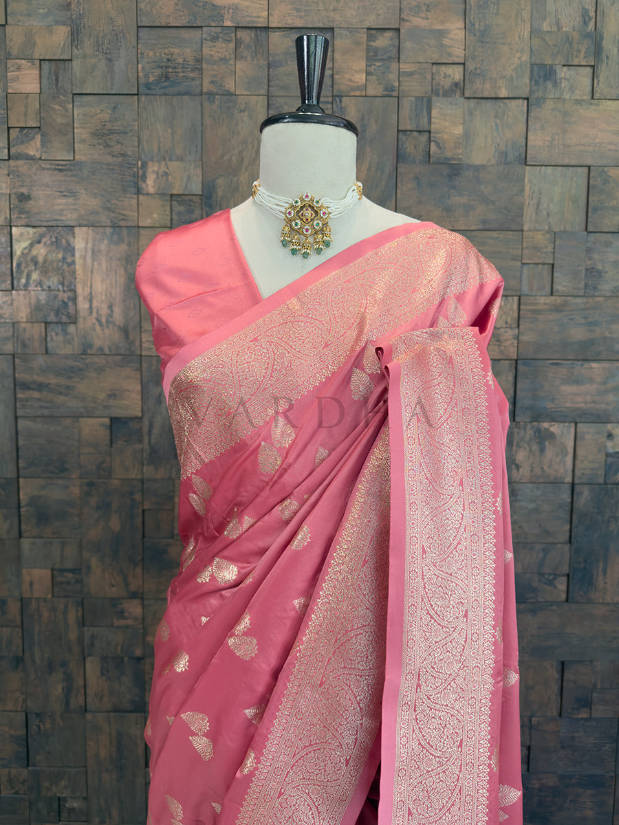 Pink saree with intricate patterns on a mannequin against a wooden paneled wall. Close-up of a soft pink woven satin ethnic saree styled on a mannequin display, paired with a traditional choker necklace.