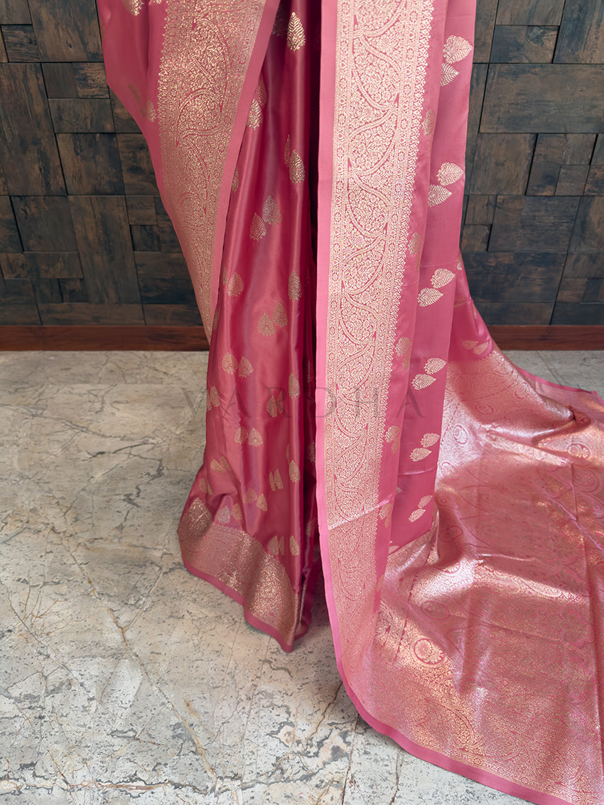 Pink saree with gold patterns on a stone floor and wooden wall background Close-up texture shot highlighting the woven leaf bootis and dense floral zari border on a dusty rose pink satin fabric. Elegant party wear rose pink satin silk saree highlighting its smooth drape, lustrous shine, and intricate zari craftsmanship.