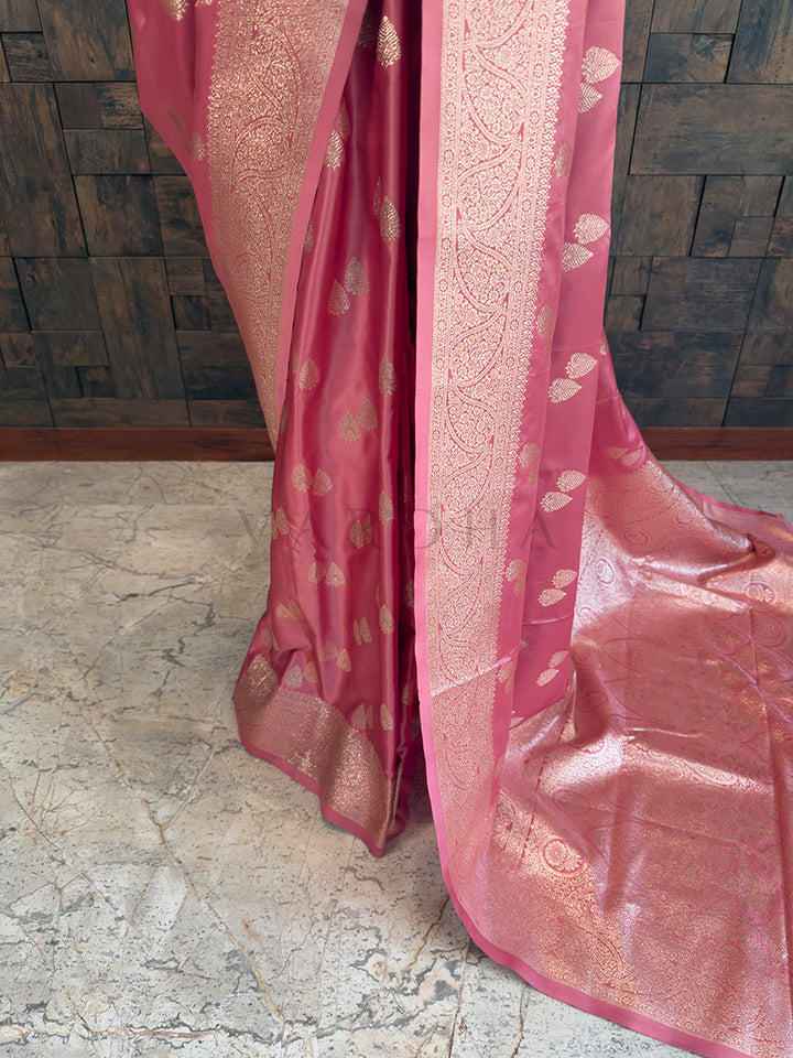 Pink saree with gold patterns on a stone floor and wooden wall background Close-up texture shot highlighting the woven leaf bootis and dense floral zari border on a dusty rose pink satin fabric. Elegant party wear rose pink satin silk saree highlighting its smooth drape, lustrous shine, and intricate zari craftsmanship.