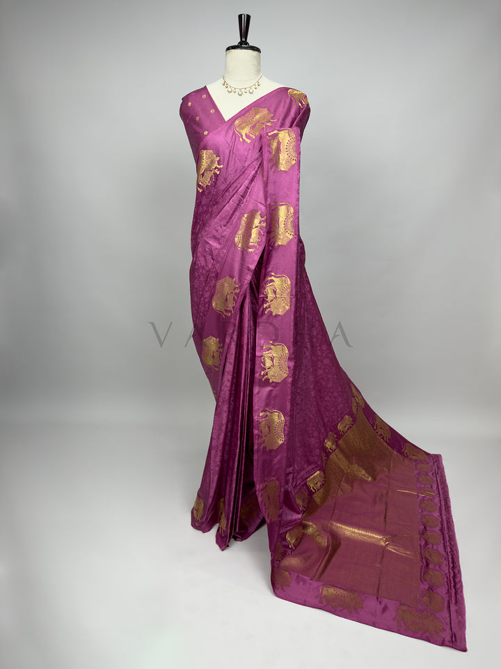 A full-length presentation of a Dusty Rose Satin Silk Saree elegantly draped on a mannequin against a neutral gray background. The saree features a soft, sophisticated dusty rose base enhanced with a subtle self-jacquard floral texture, beautifully complemented by large gold zari elephant motifs woven across the drape. The heavy, gold-woven pallu flows fluidly onto the floor, showcasing a luxurious and graceful festive ensemble. Purple saree with gold patterns on a mannequin against a gray background