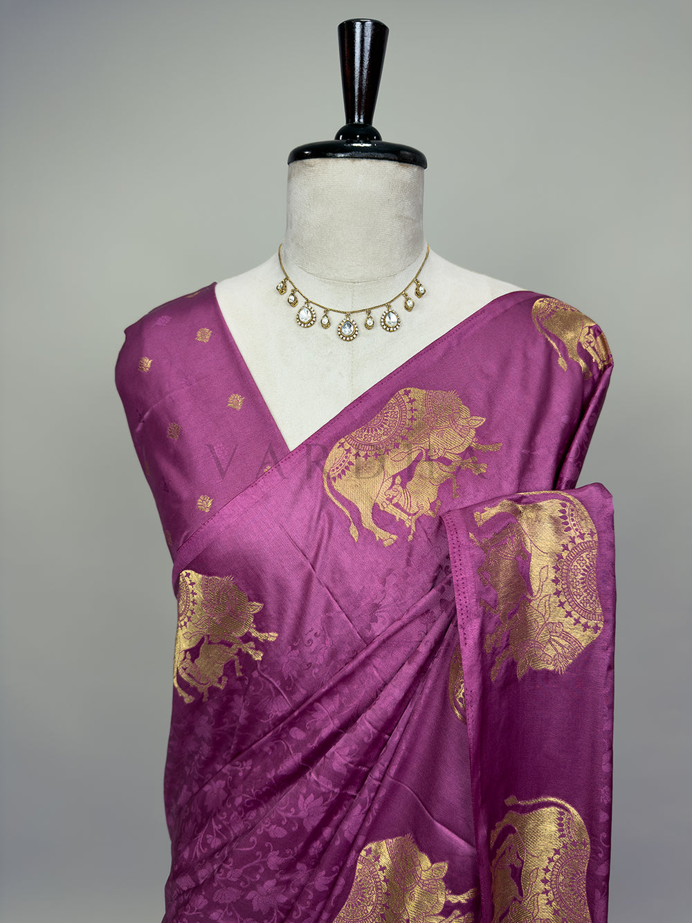 A detailed half-torso perspective of the draped Dusty Rose Satin Silk Saree on a mannequin, styled with a classic delicate stone necklace. This angle highlights the exquisite pattern layout over the chest and pleated shoulder drape, emphasizing the embossed tone-on-tone floral vine designs and the intricately woven gold zari elephant motifs against the glossy satin surface. Purple saree with gold patterns on a mannequin against a gray background