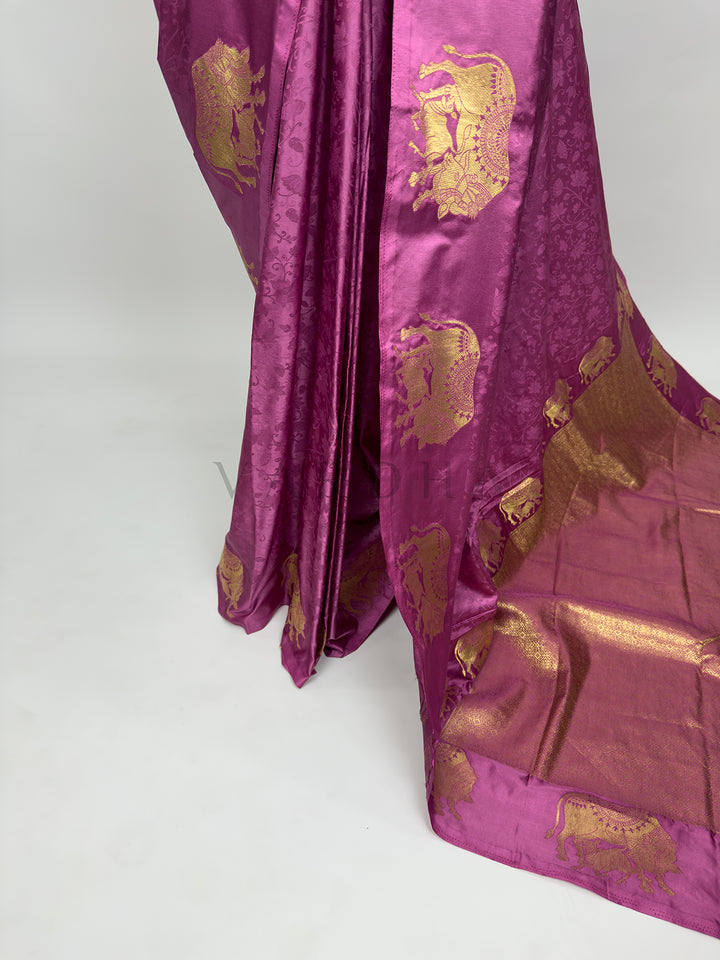 A targeted shot focusing on the lower drape and front pleats of the Dusty Rose Satin Silk Saree. The image captures the smooth, reflective sheen of the premium satin fabric as it forms structured, clean vertical folds. A prominent vertical panel decorated with a repeating sequence of detailed gold zari elephant motifs frames the side, flowing seamlessly into the grand pallu. Purple fabric with gold patterns on a white background