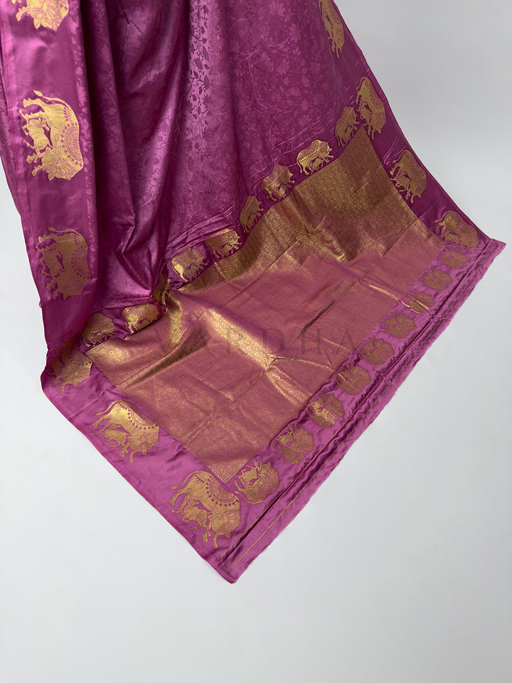 A high-angle shot showcasing the grand, fully extended pallu of the Dusty Rose Satin Silk Saree spread out across the floor. The center of the palla features a heavy, dense geometric grid pattern woven entirely from shimmering gold zari threads, which is beautifully bordered by a detailed parade of marching elephant motifs, celebrating classic Banarasi handloom heritage. Purple fabric with gold elephant patterns on a white background