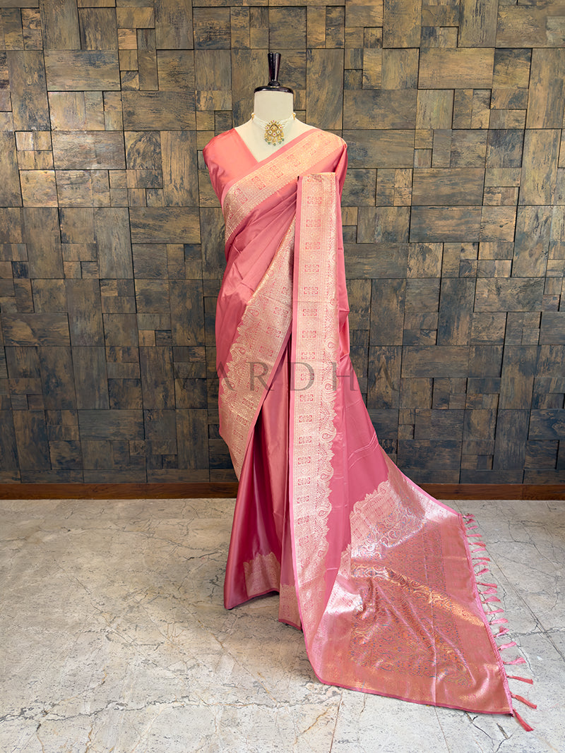 Pink saree with gold border on a mannequin against a textured wall. Full front display of a solid dusty rose pink satin silk saree featuring a heavy golden zari border draped gracefully on a mannequin.