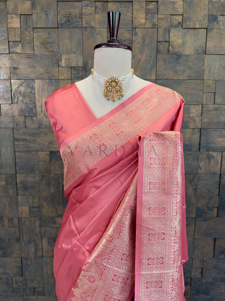Pink saree with intricate patterns on a mannequin against a textured wall. Close-up of a soft pink woven satin ethnic saree styled on a mannequin display, paired with a traditional pearl choker necklace.