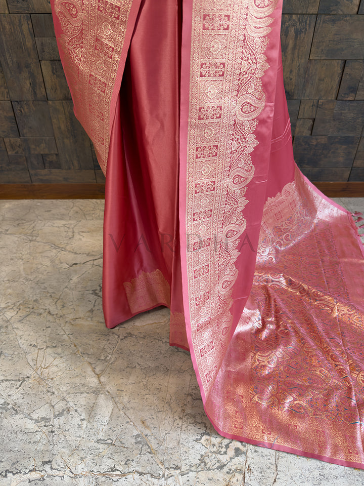 Pink saree with intricate patterns on a stone floor and wooden wall Lower pleats and fluid drape of a glossy dusty rose Banarasi-style satin silk saree showcasing the broad geometric floral border.background