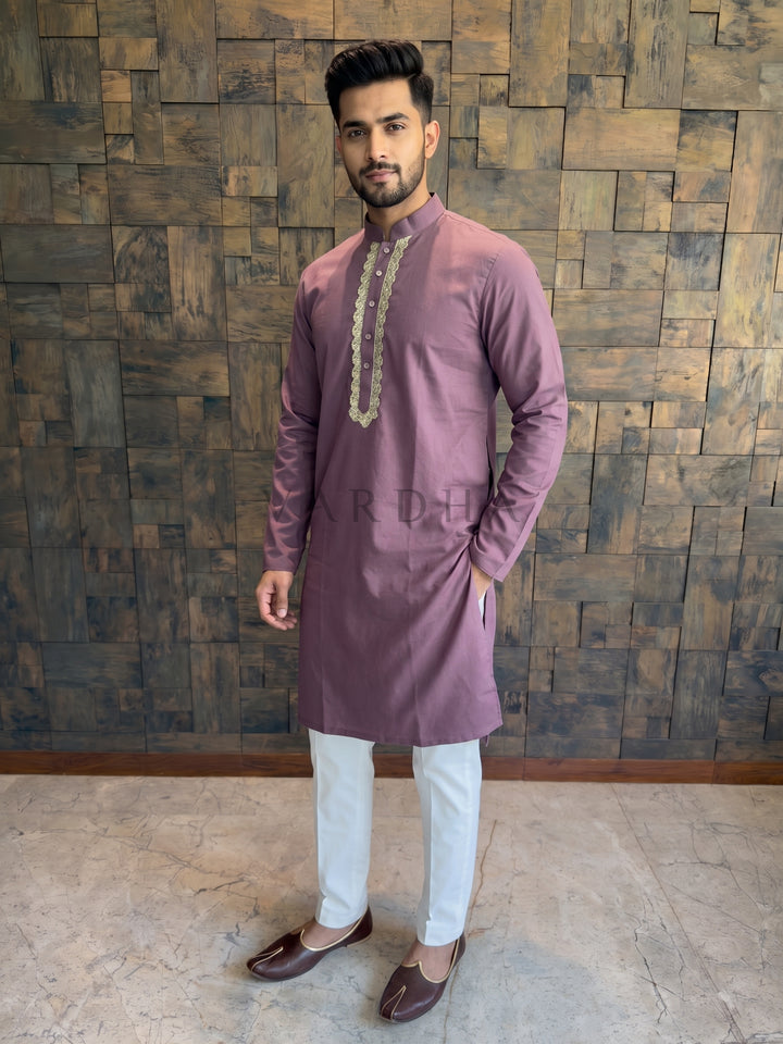 Man wearing a mauve kurta with white pants against a wooden paneled wall. Men’s Dusty Wine cotton kurta featuring a central chest placket framed by a thick, ornate scalloped gold Zari border and crystal-studded buttons.
