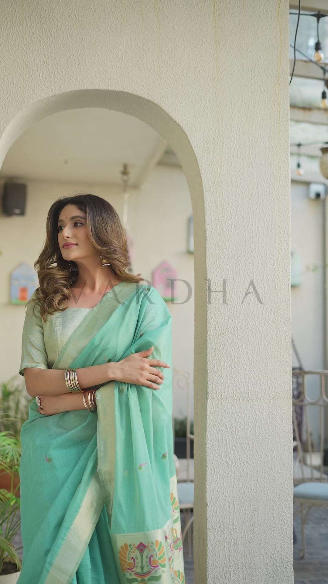 A woman wearing a mint green saree with a white border and gold embroidery, standing in front of a beige arched doorway. Graceful half-length pose of a woman looking out, highlighting the rich breathable texture, premium fabric, and regal festive appeal of the green cotton jacquard saree.