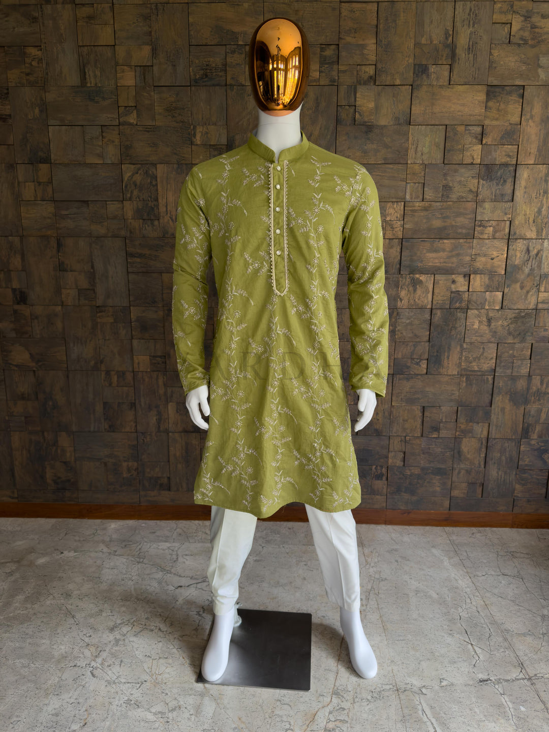 Mannequin wearing a green kurta with white patterns against a wooden paneled wall. Front view of a man wearing a regal emerald green viscose kurta featuring all-over floral vine embroidery. This is an incredibly rich, regal Emerald Green Deep Bottle Green long kurta. Dark, jewel-toned greens are traditional favorites for evening celebrations, making this piece an ideal anchor for Sangeet nights, Mehendi events, and formal wedding receptions.