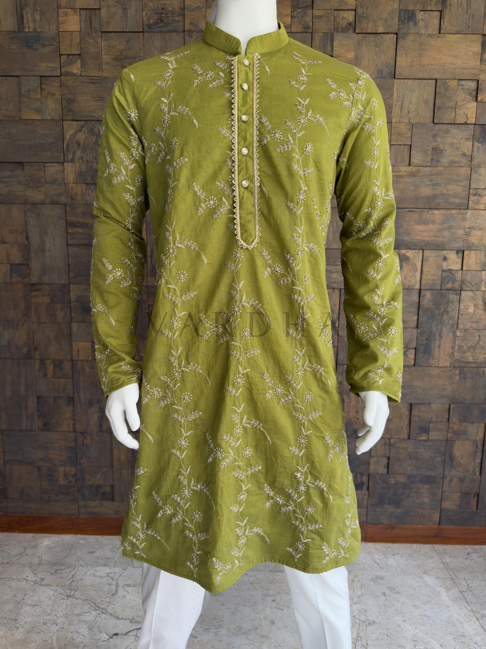 Green embroidered kurta on a mannequin against a wooden paneled wall. Close-up of the deep green kurta showcasing the intricate floral threadwork and a striking gold zig-zag bordered placket. Tailored with a clean Mandarin collar. The brilliantly embellished placket is fastened with premium, crystal-studded designer buttons, adding a final layer of evening-ready sparkle. A classic knee-length Long Kurta with full, straight sleeves and a clean, straight hem.