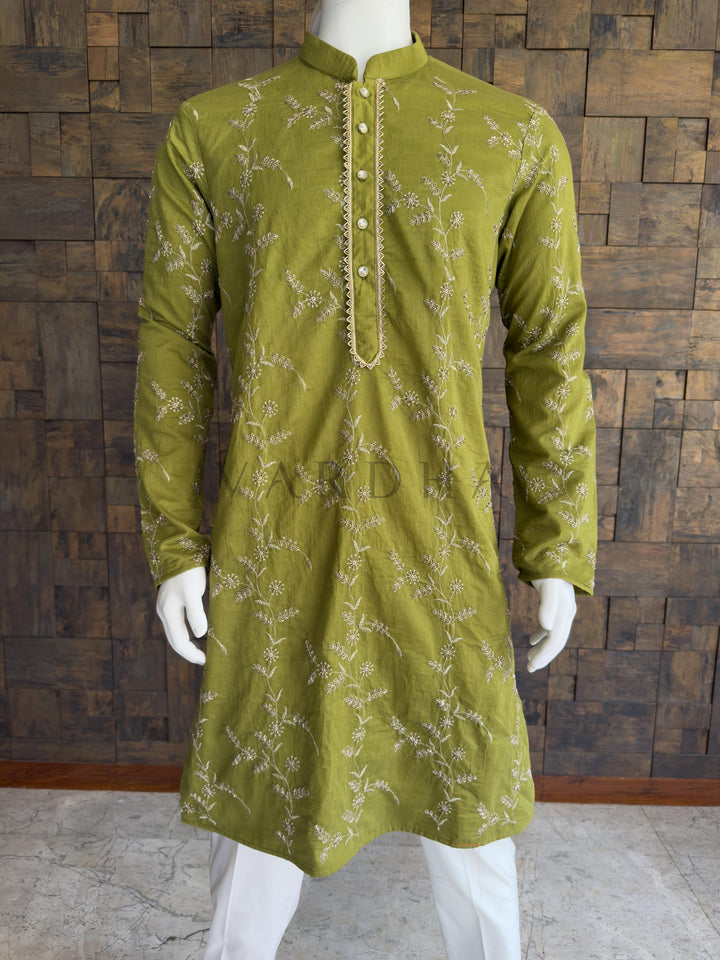 Green embroidered kurta on a mannequin against a wooden paneled wall. Close-up of the deep green kurta showcasing the intricate floral threadwork and a striking gold zig-zag bordered placket. Tailored with a clean Mandarin collar. The brilliantly embellished placket is fastened with premium, crystal-studded designer buttons, adding a final layer of evening-ready sparkle. A classic knee-length Long Kurta with full, straight sleeves and a clean, straight hem.