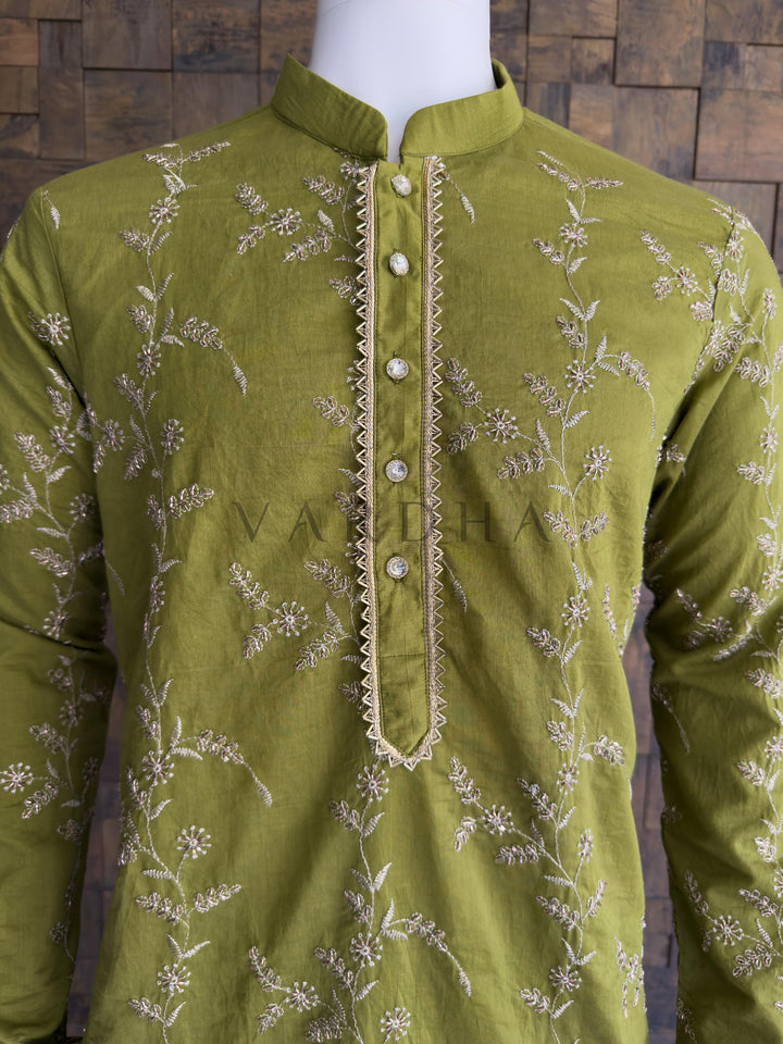 Green embroidered shirt on a mannequin against a wooden paneled wall. Detailed view of the crystal-studded designer buttons and geometric gold zari border on the emerald festive kurta. This fabric provides a luxurious, silk-like drape and a beautiful natural sheen. It flows elegantly over the body, carrying the heavy all-over embroidery perfectly without feeling stiff or restrictive.