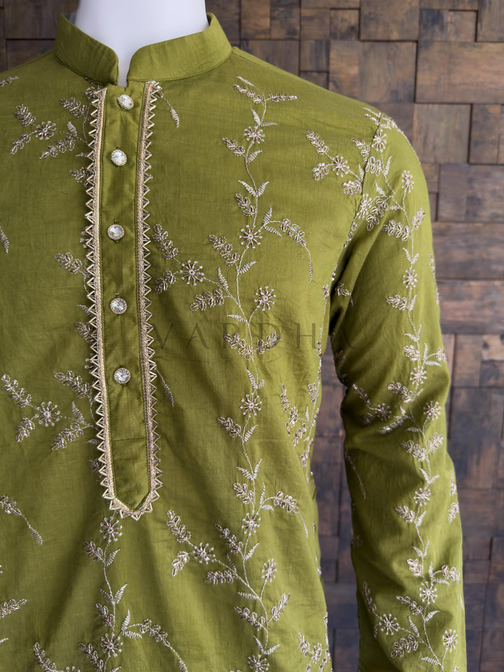 Green embroidered shirt on a mannequin against a wooden paneled wall. Side profile of the dark green embroidered viscose kurta for men, highlighting the luxurious, silk-like drape. This garment features exquisite, all-over delicate floral and vine embroidery. The intricate threadwork catches the light beautifully, giving the rich green base a heavily textured, premium look. The center placket provides a sharp, structured contrast with its bold gold zig-zag (chevron) zari border.