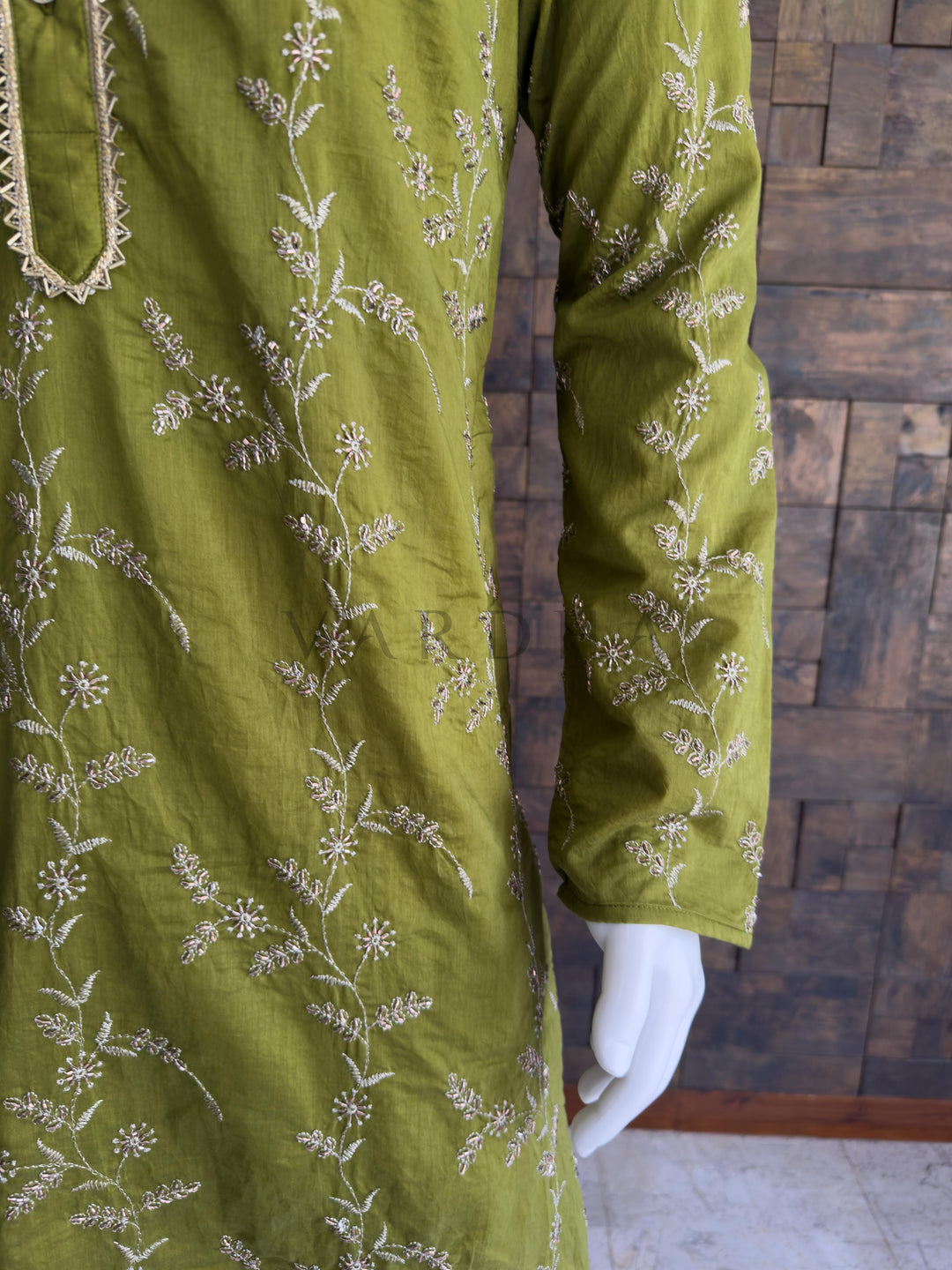 Green embroidered garment on a mannequin against a wooden paneled wall. Close-up of the full sleeve showing detailed all-over floral embroidery on the vibrant emerald green viscose fabric. This is an incredibly rich, regal Emerald Green / Deep Bottle Green long kurta. Dark, jewel-toned greens are traditional favorites for evening celebrations, making this piece an ideal anchor for Sangeet nights, Mehendi events, and formal wedding receptions.