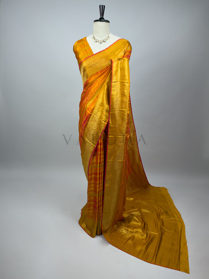 A full-length drape of an Exquisite Yellow Tanchoi Silk Saree on a mannequin adorned with a classic kundan necklace. The saree showcases rich, intricate thread weaving with dense golden-yellow textured patterns seamlessly combined with subtle orange and pink highlights along the borders. The heavy pallu cascades elegantly to the floor against a clean gray background, highlighting the luxurious weight and festive look of this bridal ethnic weGold saree with red border on a mannequin against a gray background