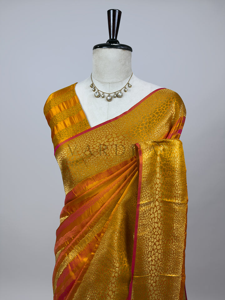 A detailed half-torso view of the draped Yellow Tanchoi Silk Saree on a mannequin, highlighting the exquisite pattern layout across the chest and shoulder drape. The intricate gold and yellow weaving creates a textured, metallic finish, accentuated by vibrant pink borders. A traditional kundan statement necklace complements the ensemble, showing how the luxury fabric frames a formal wedding look. Mannequin wearing a gold saree with red border on a gray background