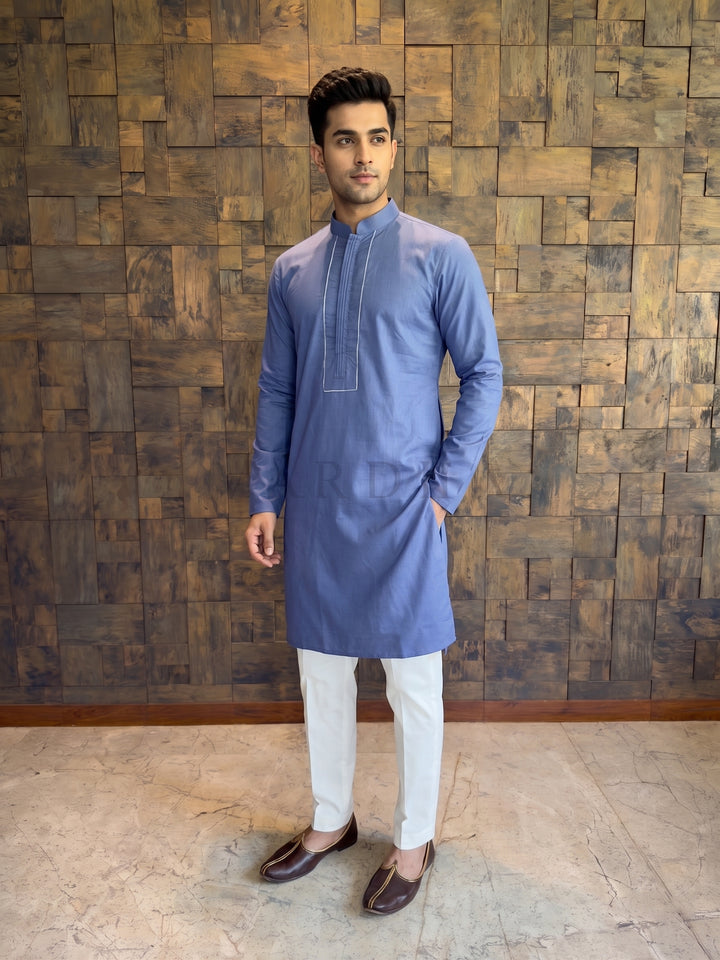 Man wearing a blue kurta with white pants against a wooden paneled wall. Front view of a French Blue cotton kurta for men, featuring a structured chest design with vertical pintucks and a crisp Mandarin collar.