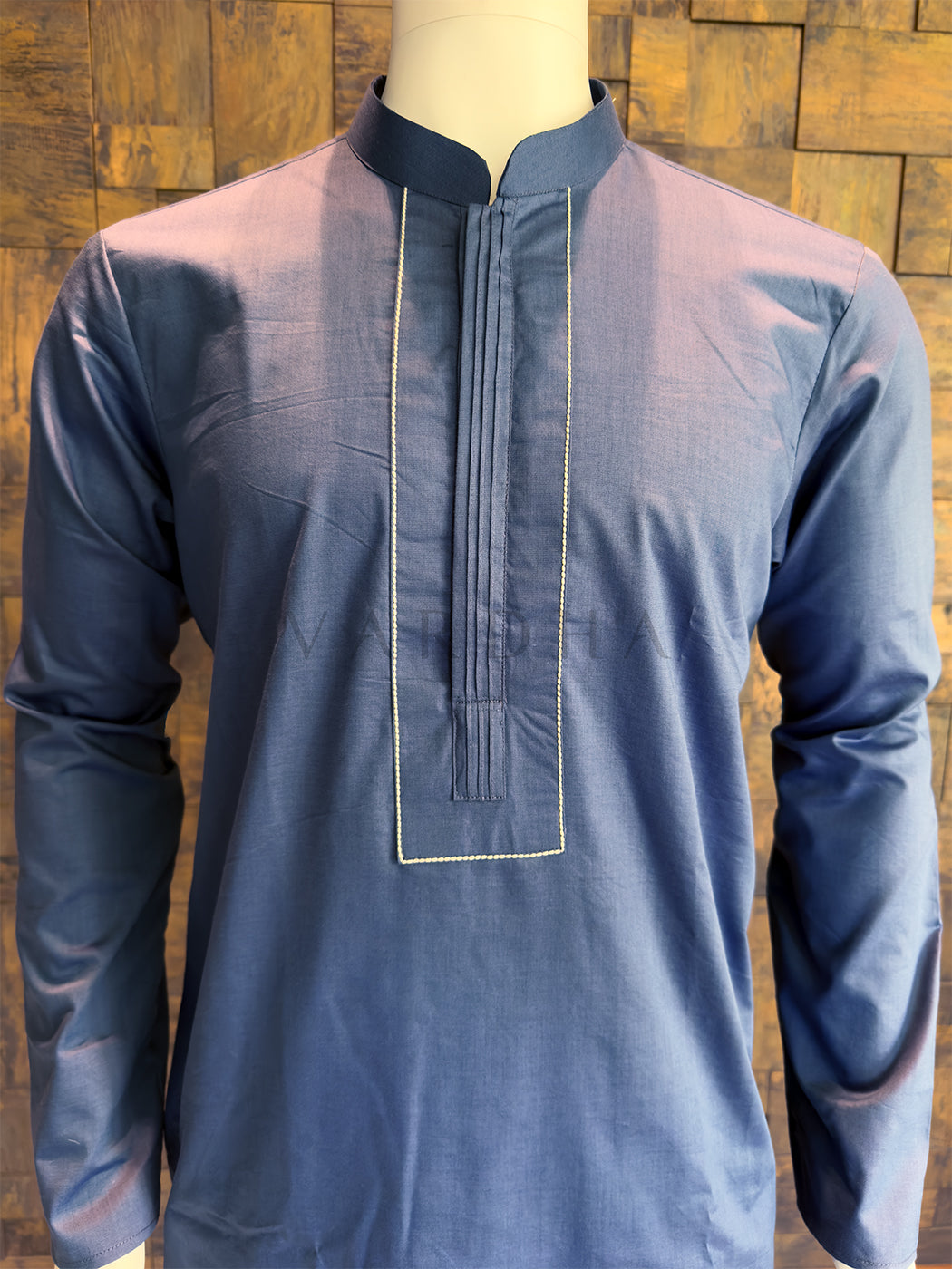 Blue long-sleeve shirt on a mannequin against a wooden paneled background. Elegant men’s ethnic wear: A knee-length French Blue cotton kurta with a minimalist architectural design, ideal for festive brunches or morning pujas.