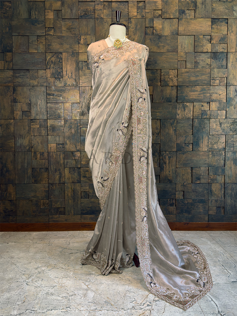 Elegant saree with intricate designs on a mannequin against a textured wall. Full View Metallic Silver Tissue Saree with Peacock Handwork, available with pre-drape services as well as free gift services
